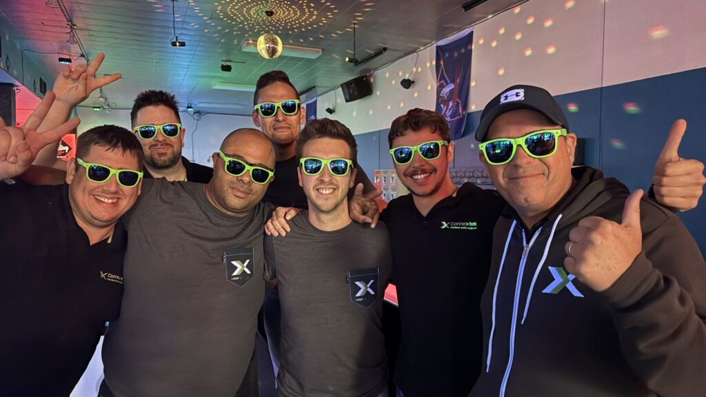 Group of men wearing neon-green sunglasses posing together indoors, smiling and giving thumbs up or peace signs, with matching X logos on shirts/farmer badges visible.